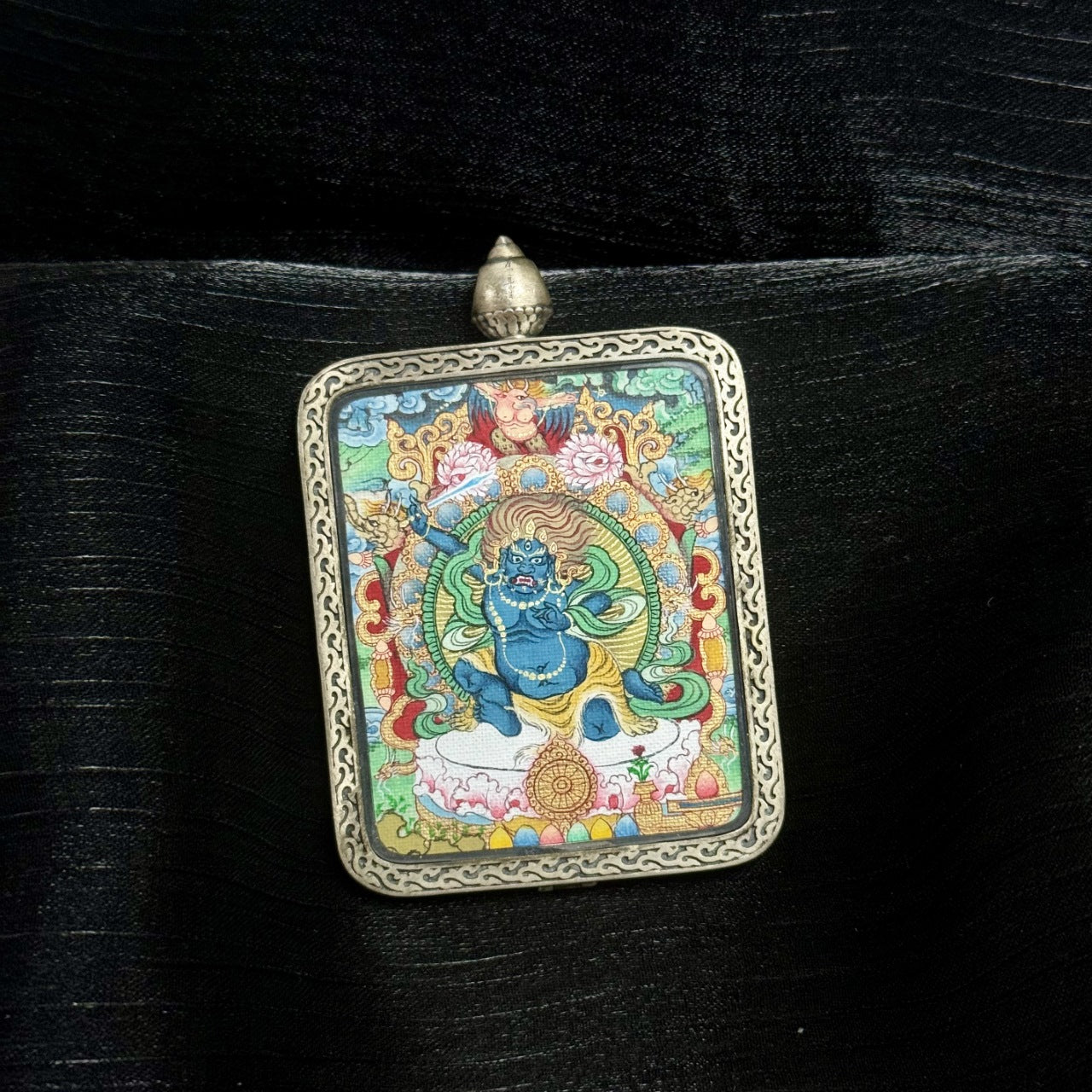 Angled close-up of Acalanatha miniature thangka pendant highlighting raised sword, dynamic posture, blue wrathful expression, layered mineral pigments, and carved silver gawu frame.