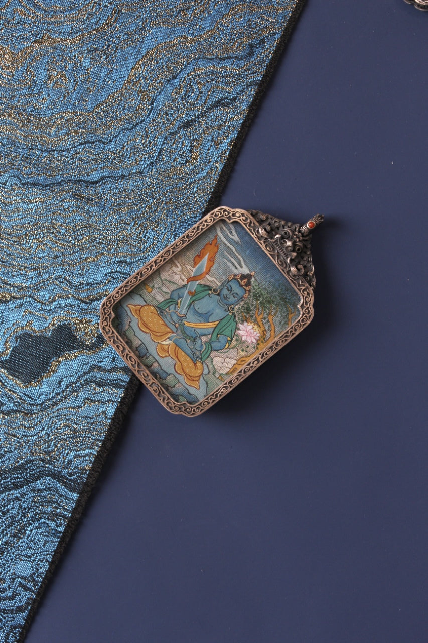 Angled flat lay of Akashagarbha Bodhisattva mini thangka pendant on traditional textile background, showing the hand-painted thangka artwork in a vintage silver case