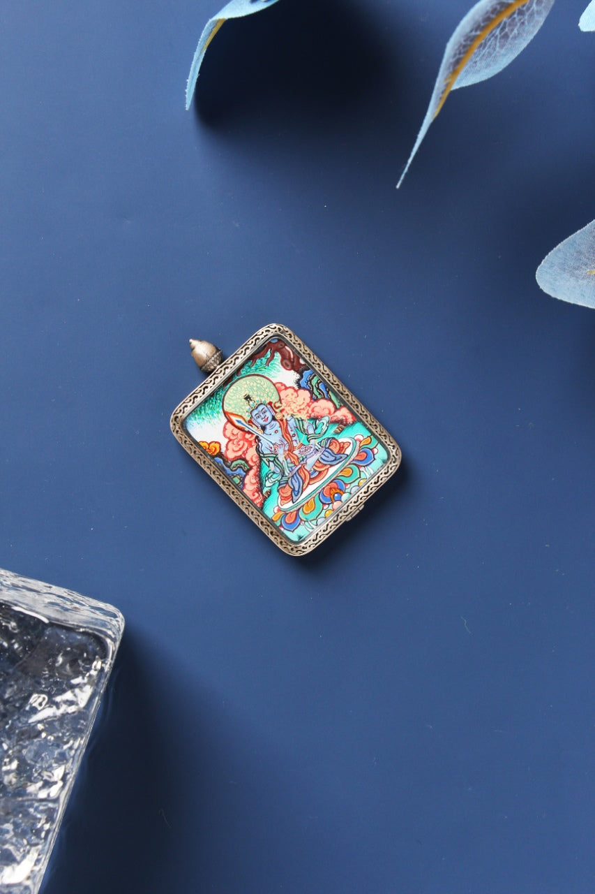 Angled flat lay of Akashagarbha Bodhisattva mini thangka pendant, hand-painted with mineral pigments and framed by a Dunhuang Nine-Color Deer-style sterling silver border, shown diagonally on a navy backdrop with translucent leaves and a clear textured stone accent.