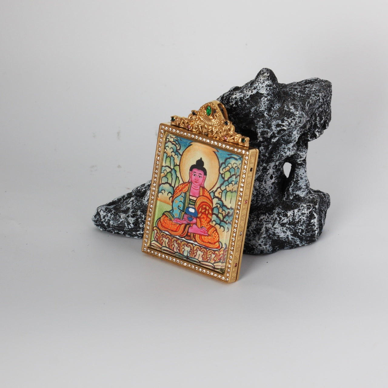 Angled view of the Amitabha Buddha mini thangka pendant highlighting the crystal-set border, gold-tone case depth, and ornate crown top with green and multicolor stones, with the hand-painted artwork visible through the front window.