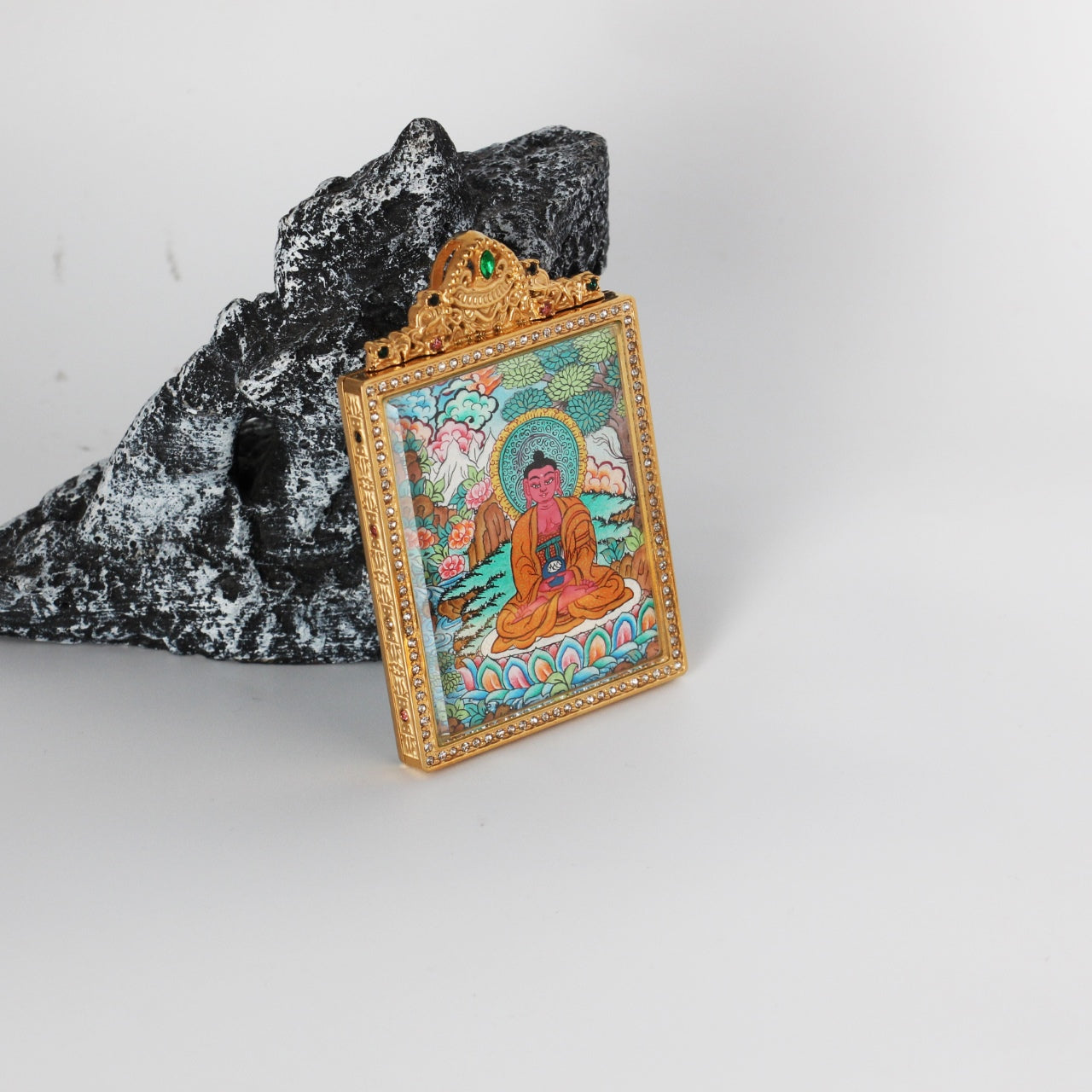 Angled view of Amitabha Buddha mini thangka pendant showing the thickness of the double-sided titanium steel case, ornate top crest with green stone detail, and vivid hand-painted thangka artwork inside