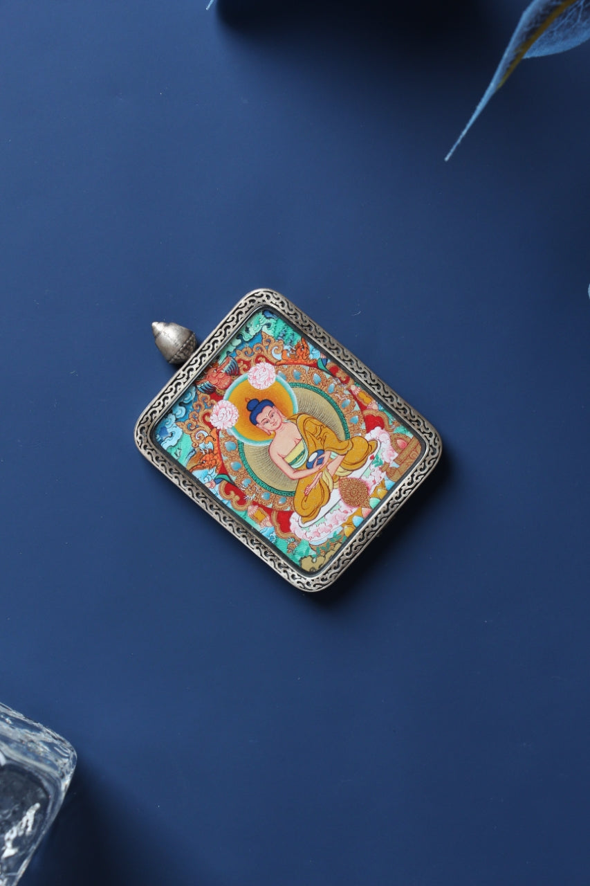 Angled view of an Amitabha Buddha mini thangka pendant with a silver gawu-style frame on a dark blue background.