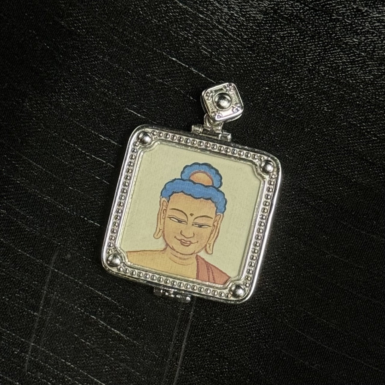 Close-up of Amitabha Buddha mini thangka pendant featuring a calm painted face with blue hair bun and soft facial lines, set in a silver-tone square frame with dotted edging, round corner beads, and a square top bail