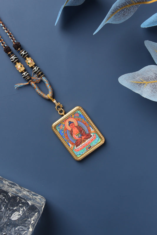 Diagonal flat-lay view of an Amitabha Buddha mini thangka pendant, featuring a hand-painted image in natural mineral pigments, set in a gold-tone titanium steel case with braided cord and bead details on a blue background.
