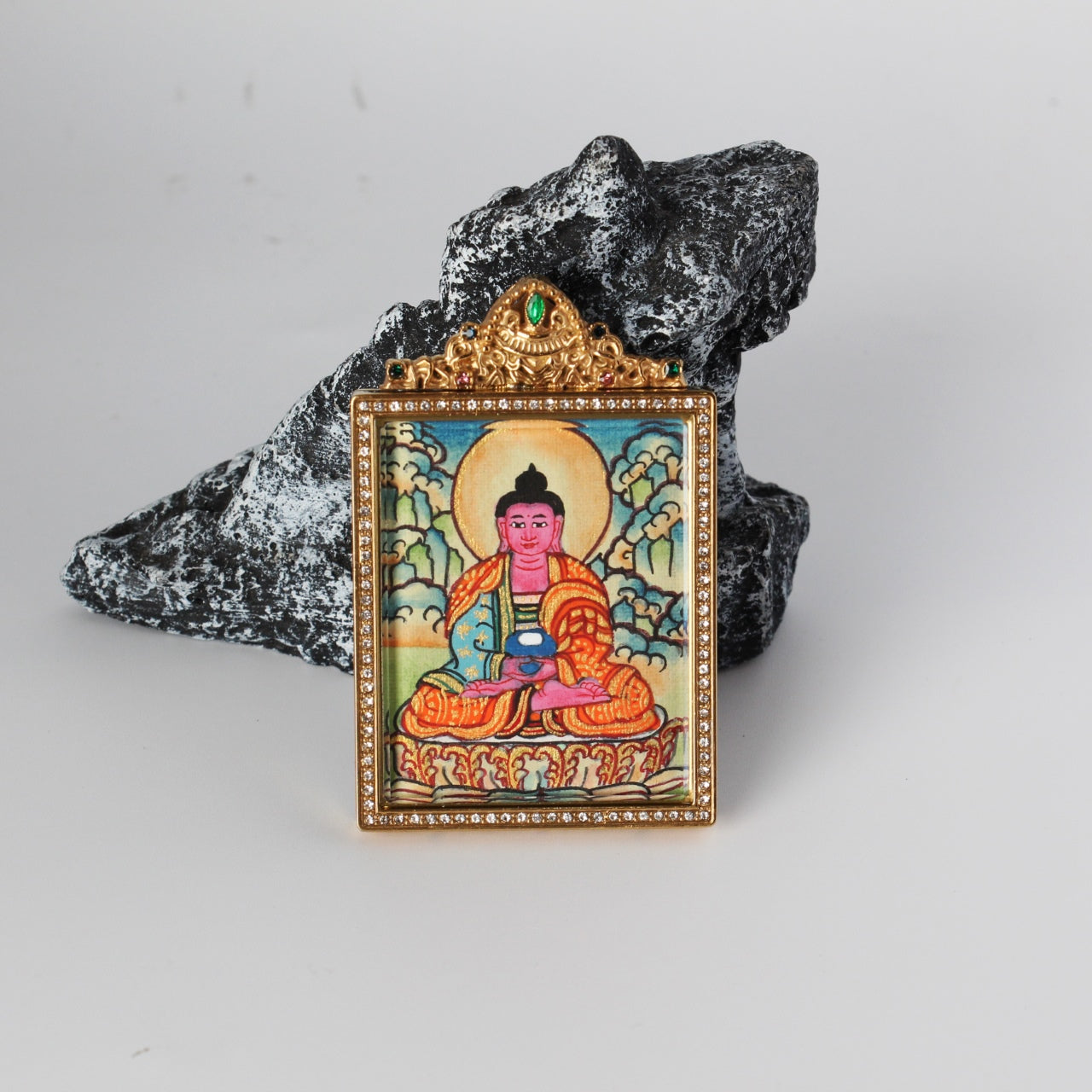 Front view of Amitabha Buddha mini thangka pendant showing the hand-painted seated Amitabha Buddha with halo, gold-tone rectangular frame with crystal border, and ornate crown top with a green gemstone, photographed on a rock prop.