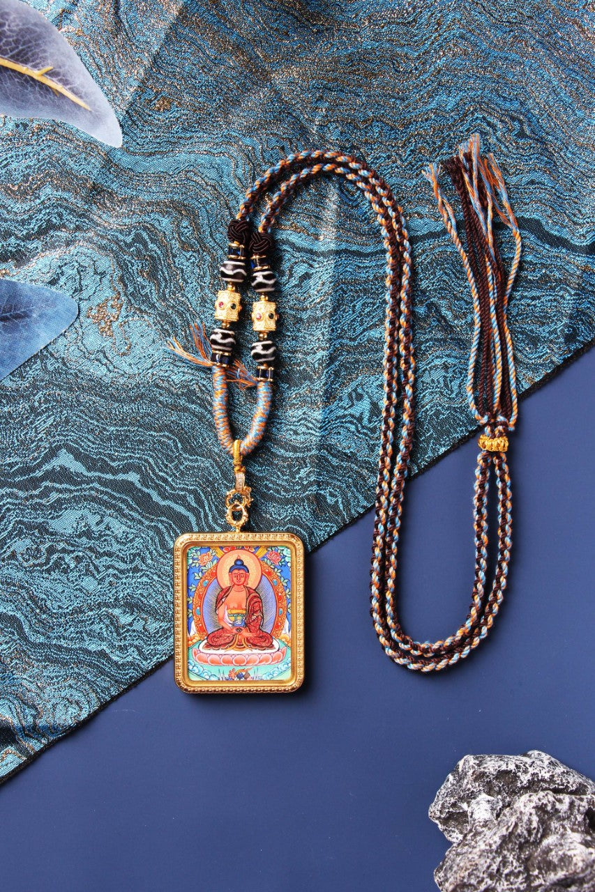 Full layout photo showing an Amitabha Buddha mini thangka pendant in a gold-tone titanium steel case, presented with a long braided cord, adjustable tassel section, and decorative bead elements.