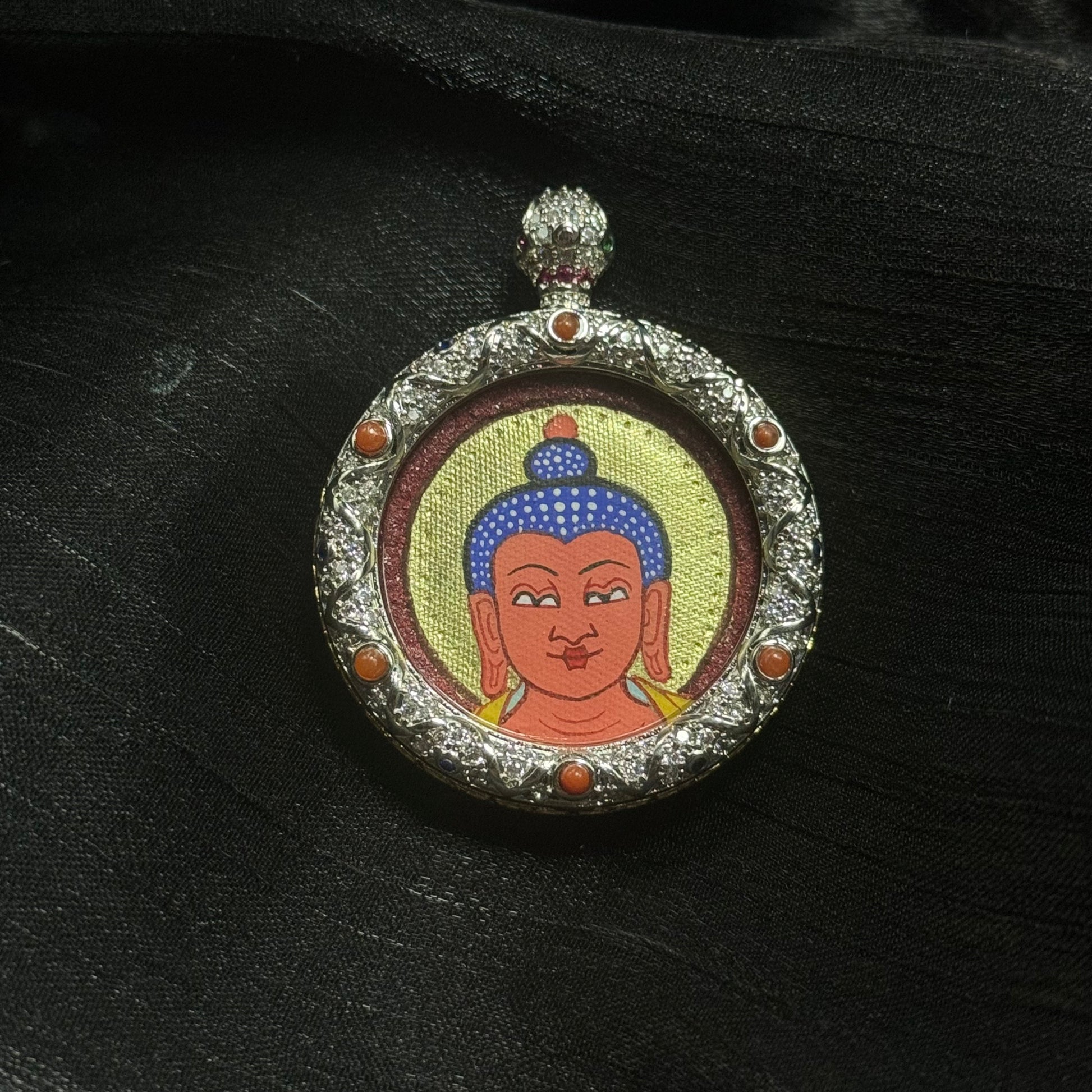 Close-up of Amitabha Buddha miniature thangka pendant showing hand-painted red Buddha head with gold halo and cinnabar mineral pigment background inside ornate jeweled copper pendant frame