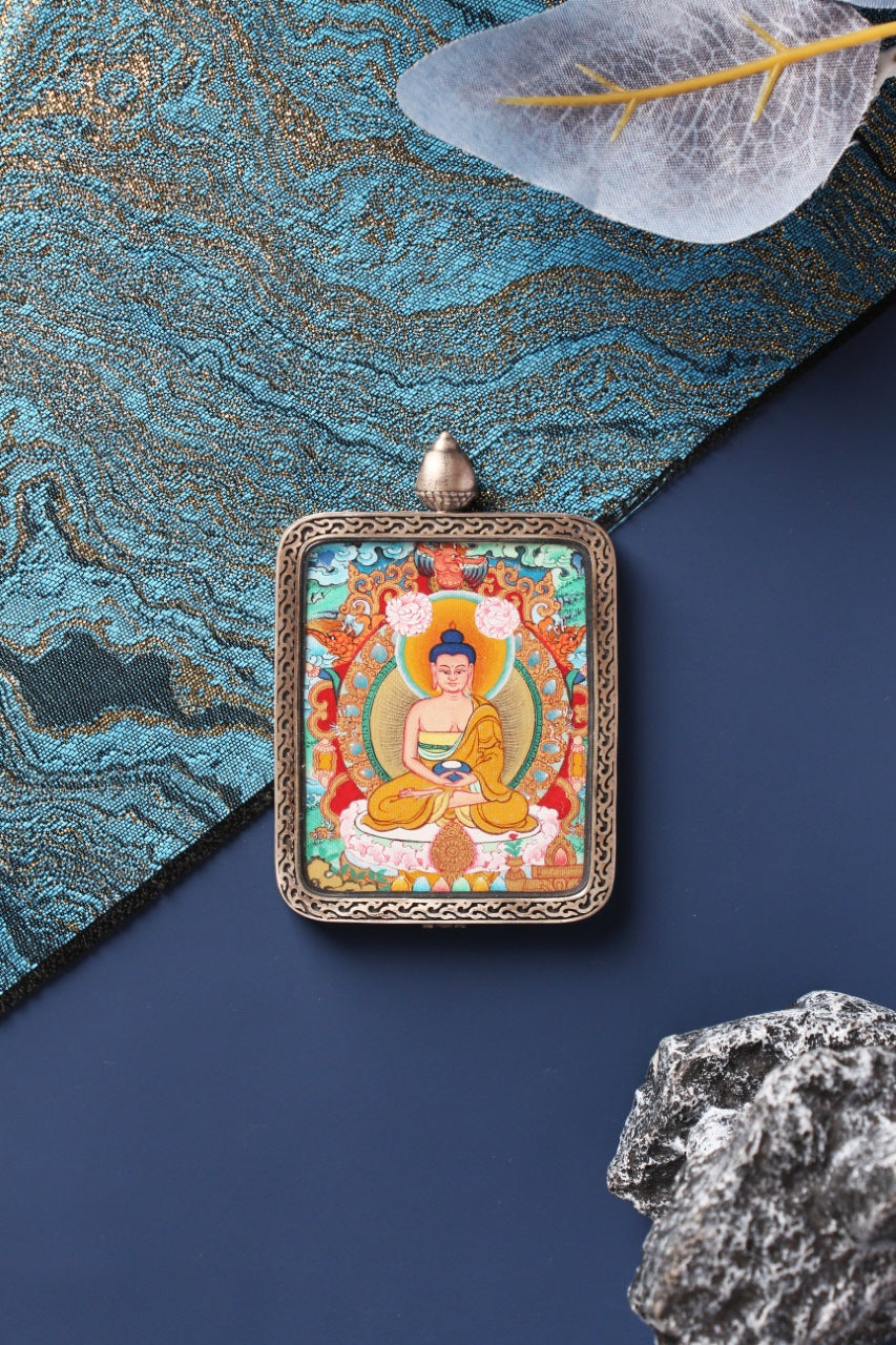 Front-facing Amitabha Buddha mini thangka pendant in a silver gawu case, displayed with blue textile and stone props.