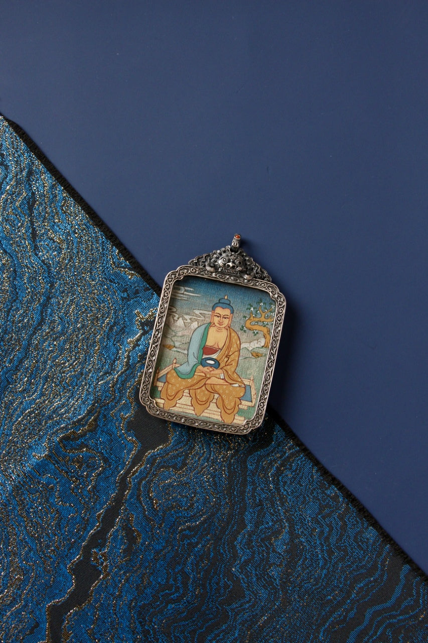 Vintage silver gawu pendant featuring a hand-painted Amitabha Buddha mini thangka, shown on a blue backdrop with brocade fabric.