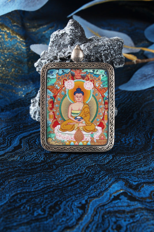 Close-up of an Amitabha Buddha mini thangka card hand-painted in Menri style, set in a silver gawu pendant case on a blue textile background.