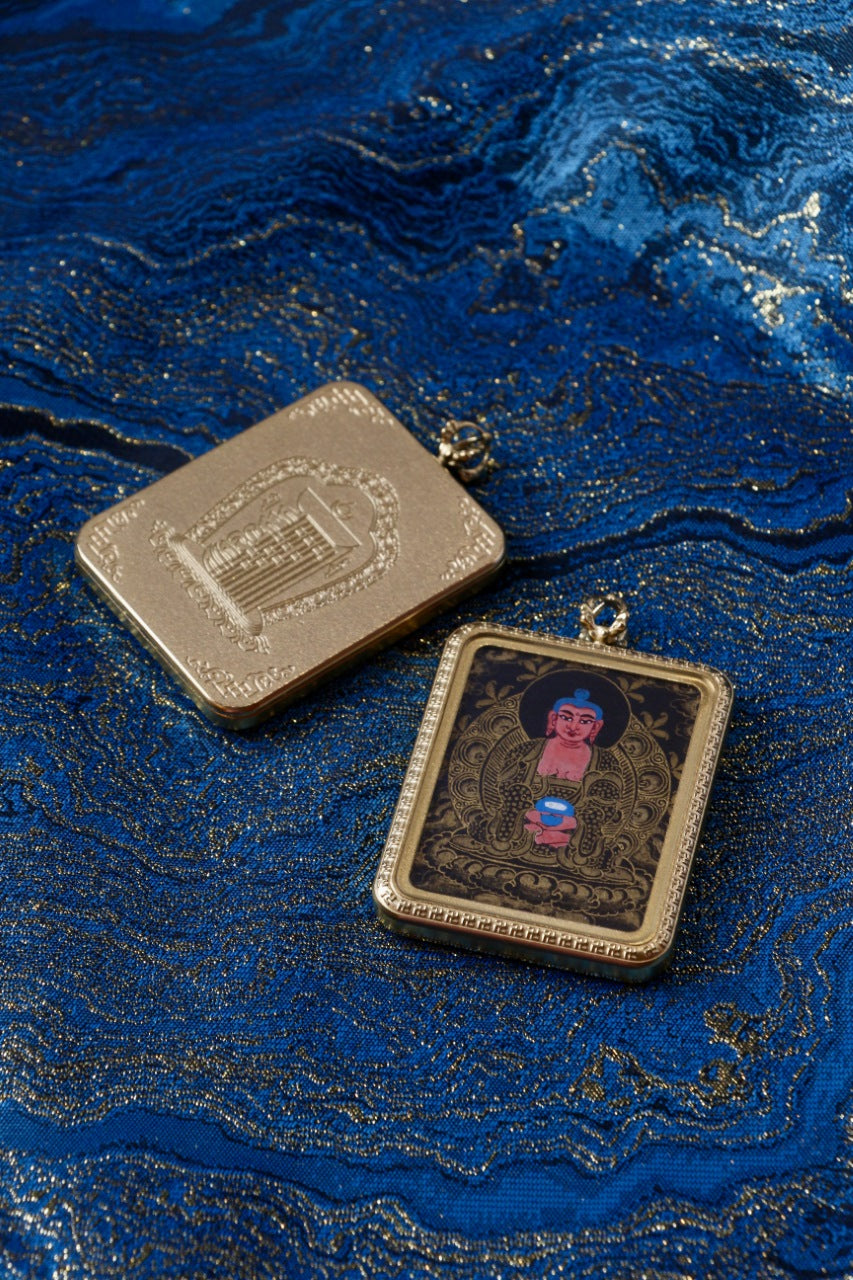 Amitabha Buddha mini thangka pendant shown with front black-gold painting and back-side metal case detail on blue brocade fabric
