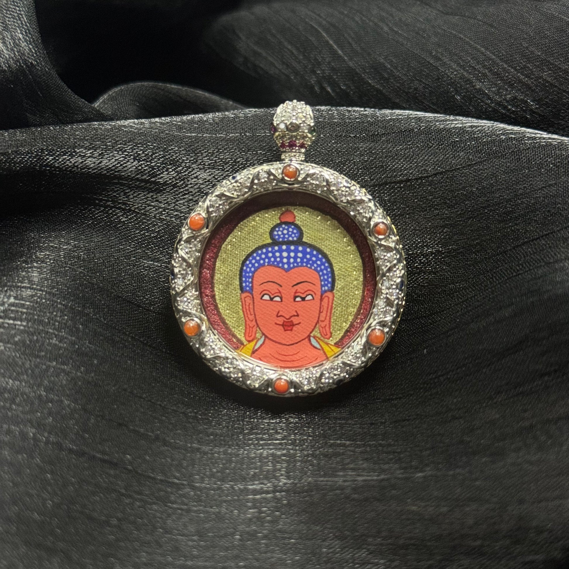 Front view of Amitabha Buddha miniature thangka pendant resting on dark silk fabric showing traditional Rebgong hand-painted Buddha head and ornate copper pendant setting