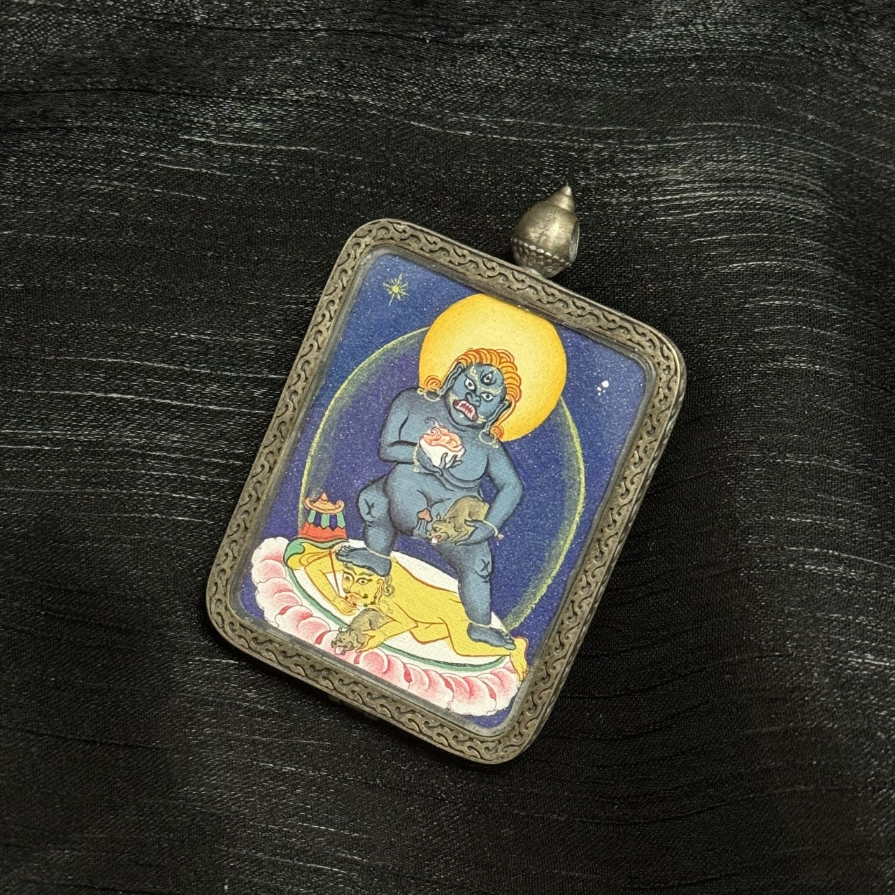 Angled view of Black Jambhala thangka pendant highlighting coarse mineral pigment texture, deep blue background, and engraved silver frame details
