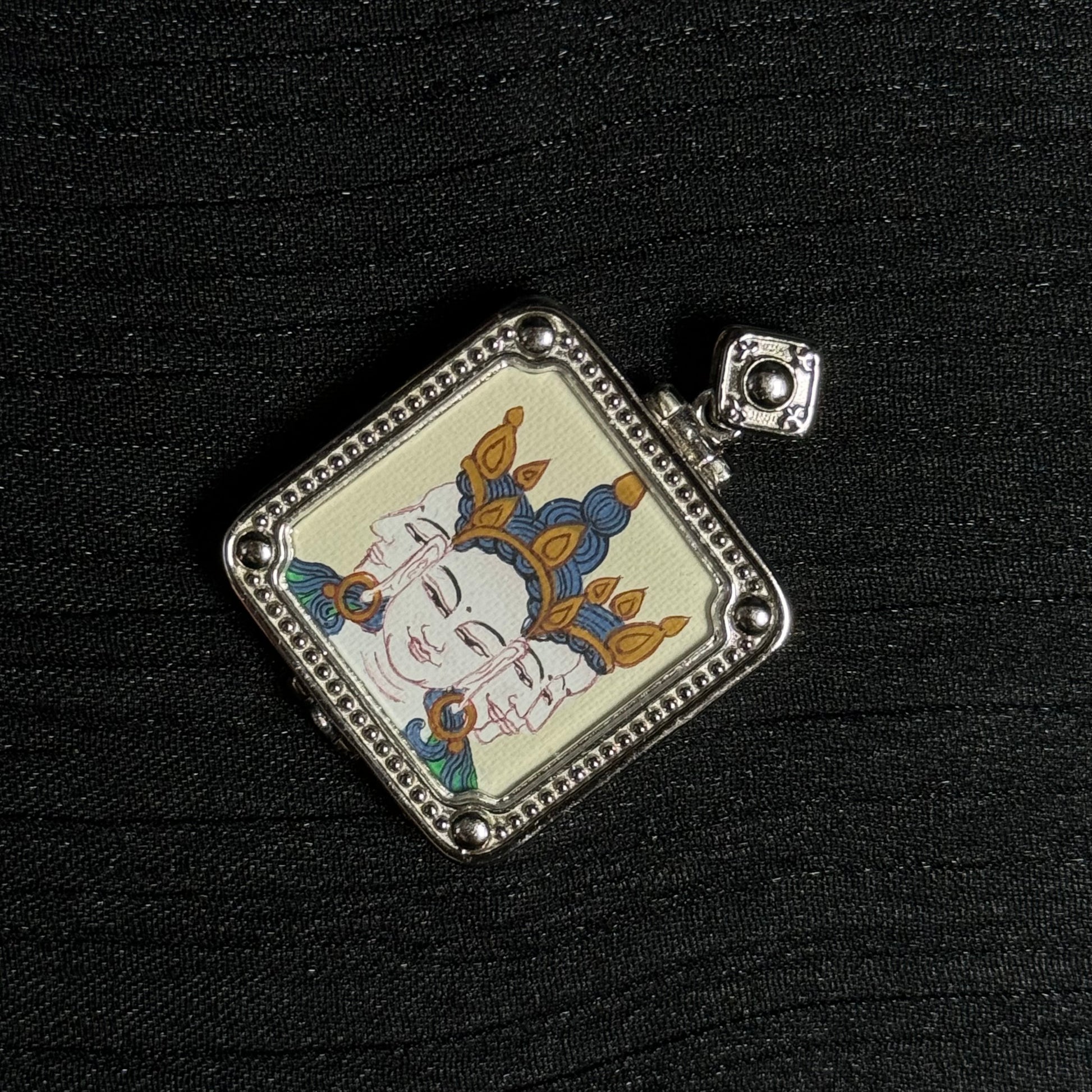 Angled view of a miniature Vairocana Buddha thangka pendant highlighting the hand-painted three-faced Buddha image and textured silver pendant frame.