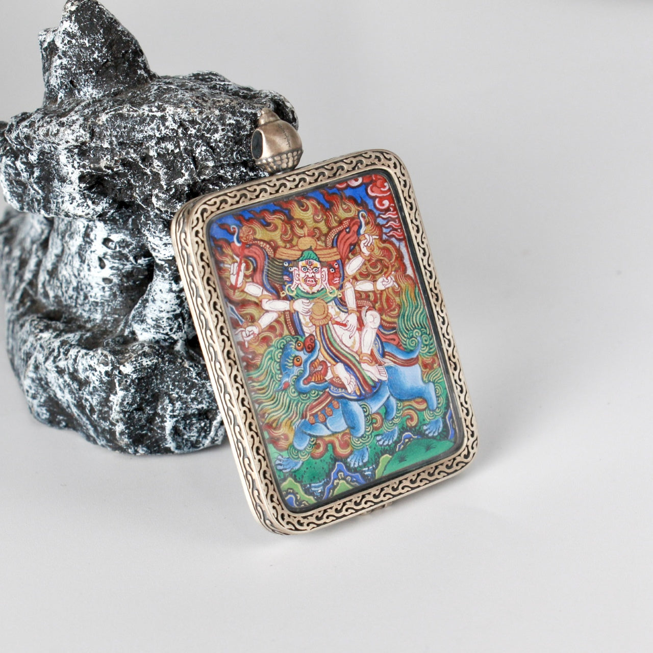 Angled view of the Baihar mini thangka pendant showing depth of mineral pigment coloration, expressive flame motifs, and the thickness and craftsmanship of the silver gawu-style case, highlighting Rebgong thangka painting characteristics.