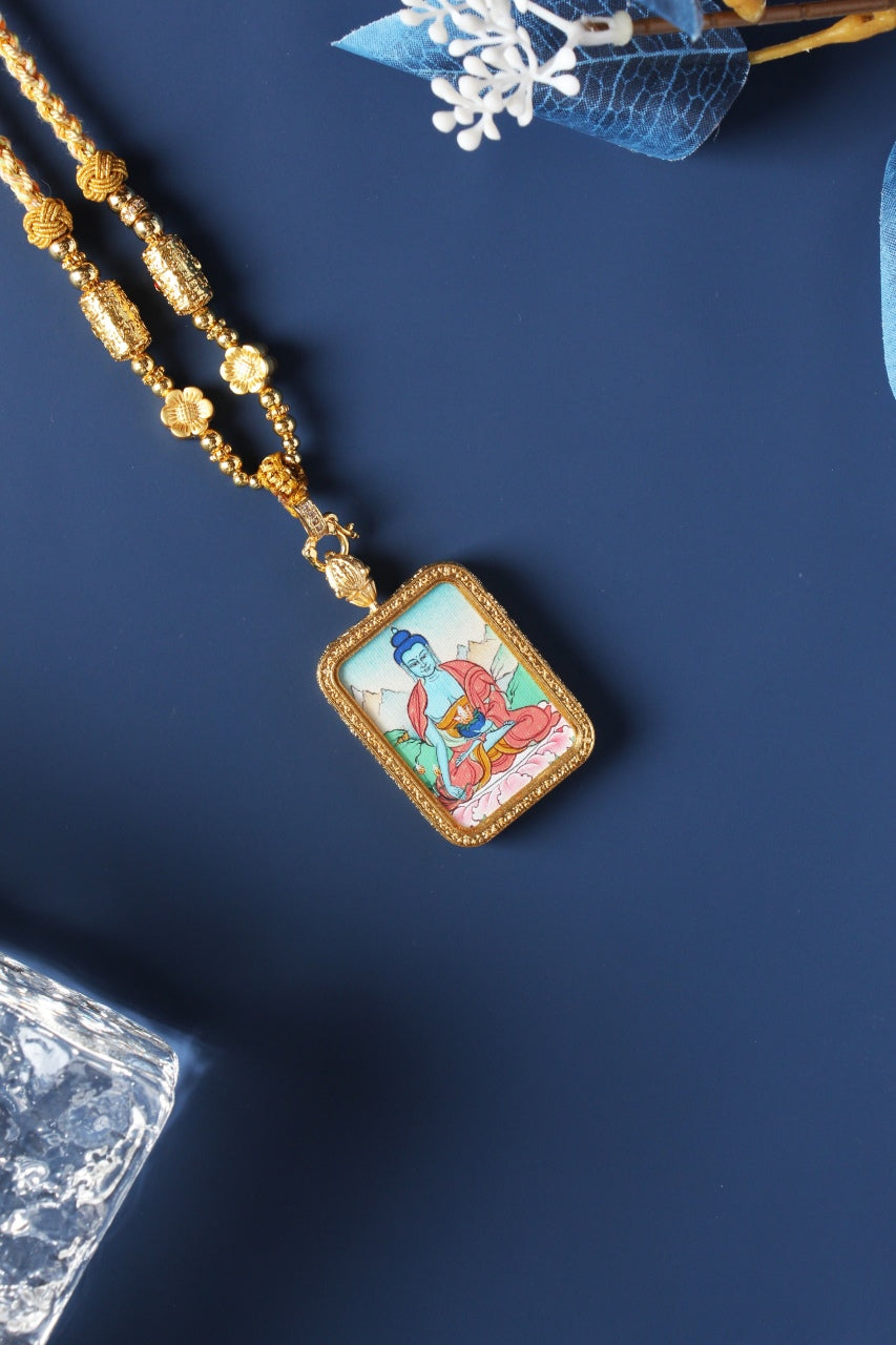 Angled close-up of a Bhaisajyaguru Buddha thangka pendant in a gold-tone frame with decorative beads.