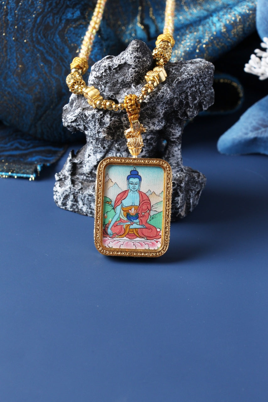 Bhaisajyaguru Buddha thangka amulet pendant standing against a stone prop, framed in gold tone with beaded necklace.