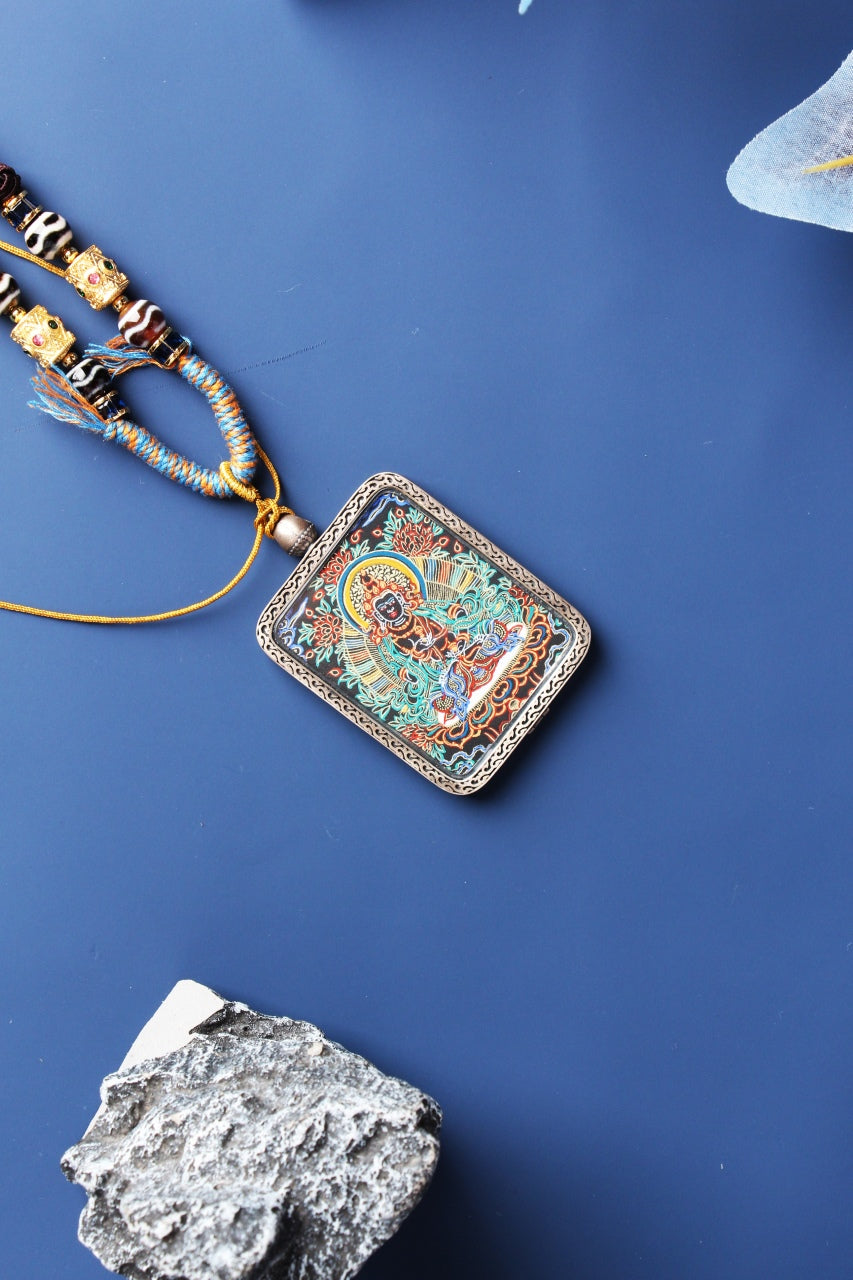 Angled flat-lay of a black-gold Vajrasattva thangka pendant with patterned cord and beadwork, highlighting the framed artwork.
