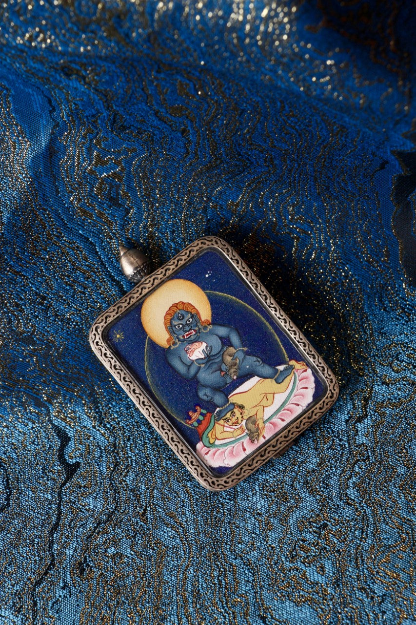 Black Jambhala miniature thangka pendant with layered mineral pigment texture, photographed on traditional blue brocade fabric