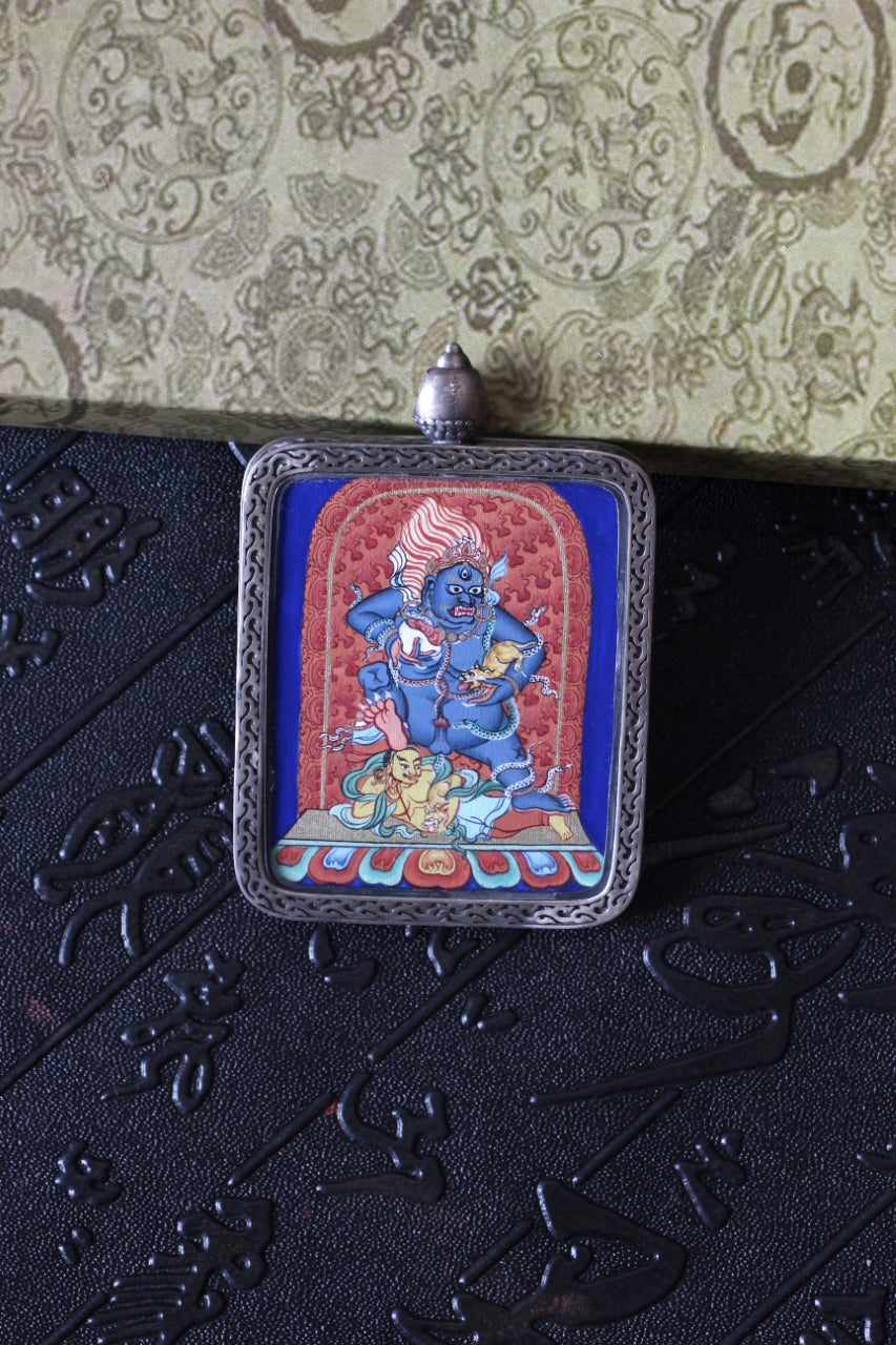 Front view of a Black Jambhala mini thangka pendant in a silver gawu case, showing vivid mineral-pigment painting and ornate silver border.