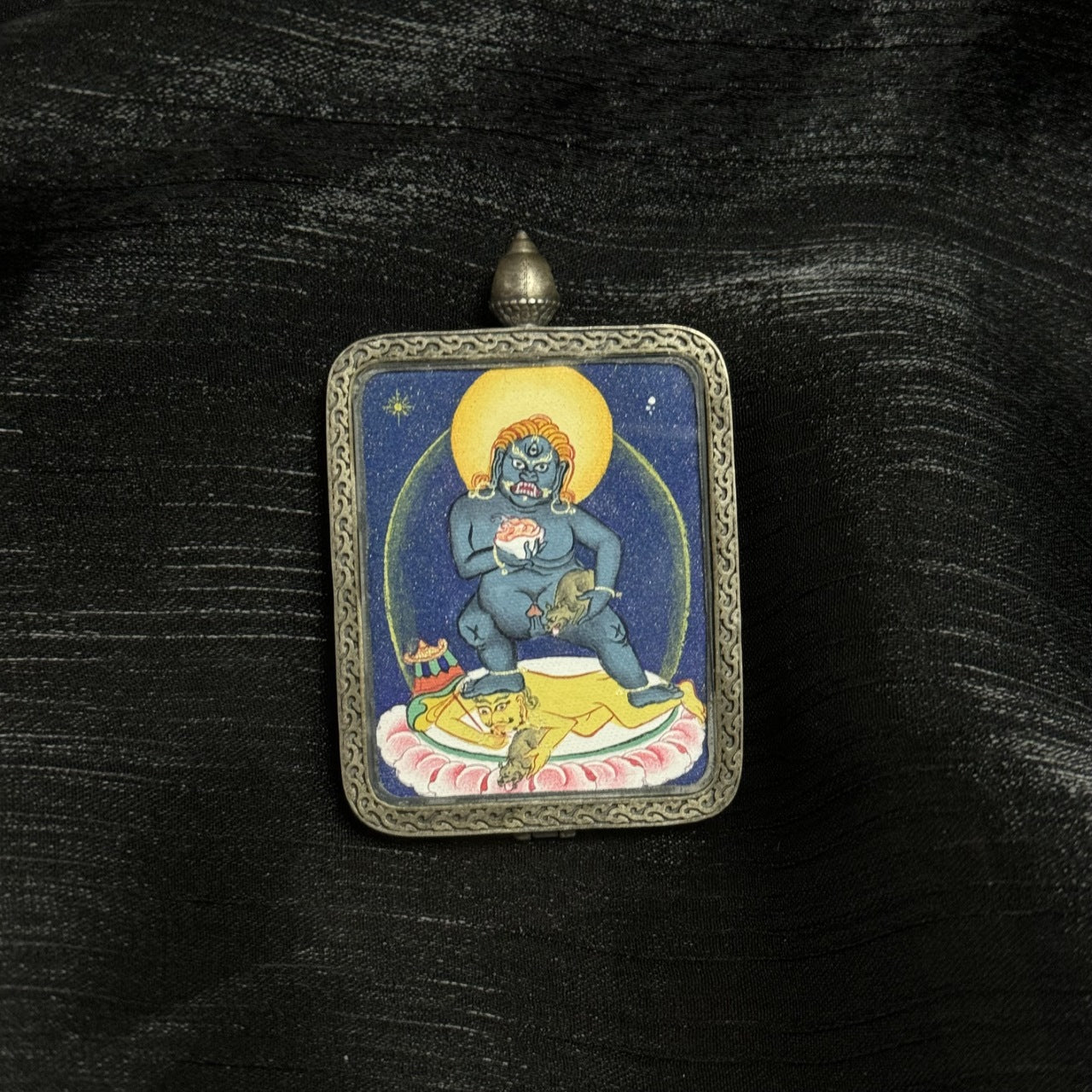 Front view of Black Jambhala mini thangka pendant, hand-painted with dense mineral pigments in Tibetan coarse mineral style, set in a traditional silver protective case