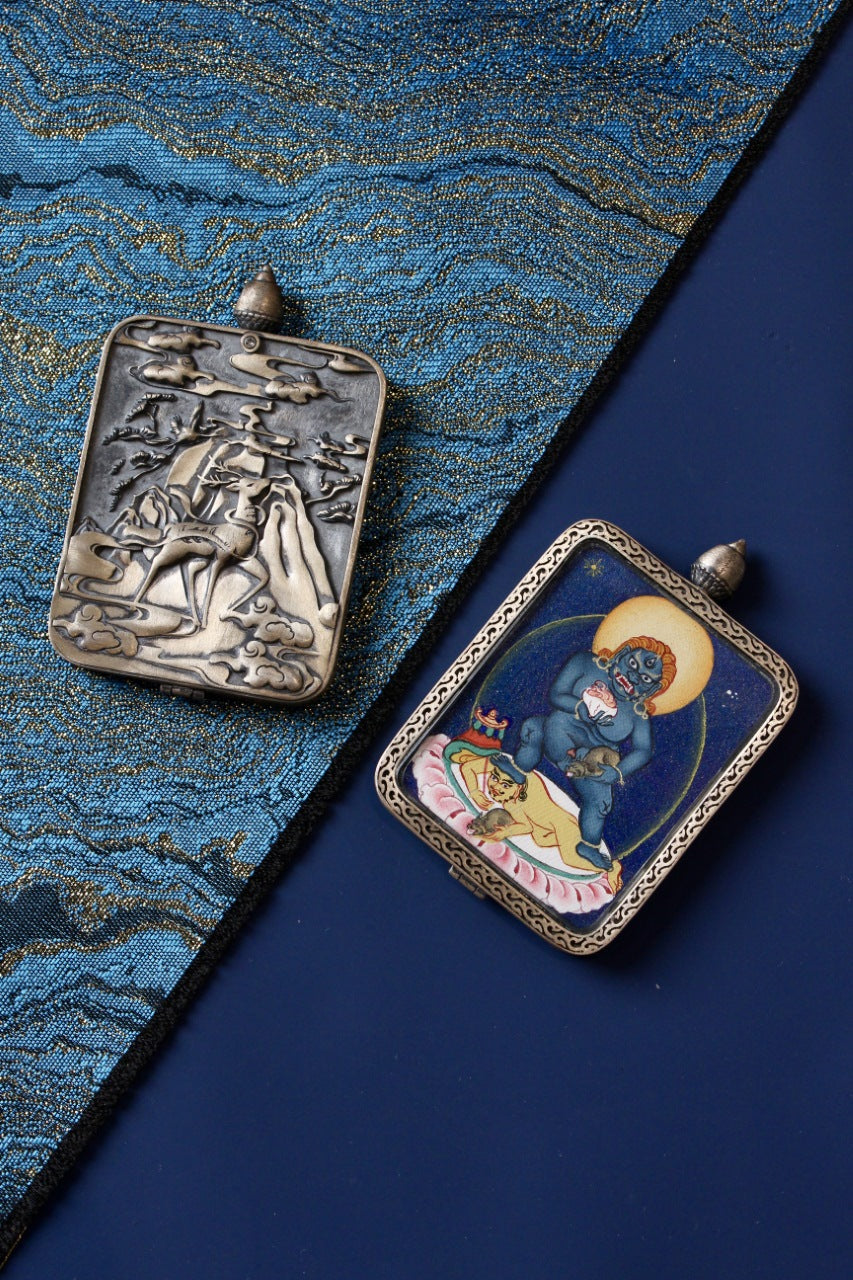 Hand-painted Black Jambhala mini thangka pendant with Tibetan mineral pigments, shown in a silver protective case on blue brocade fabric