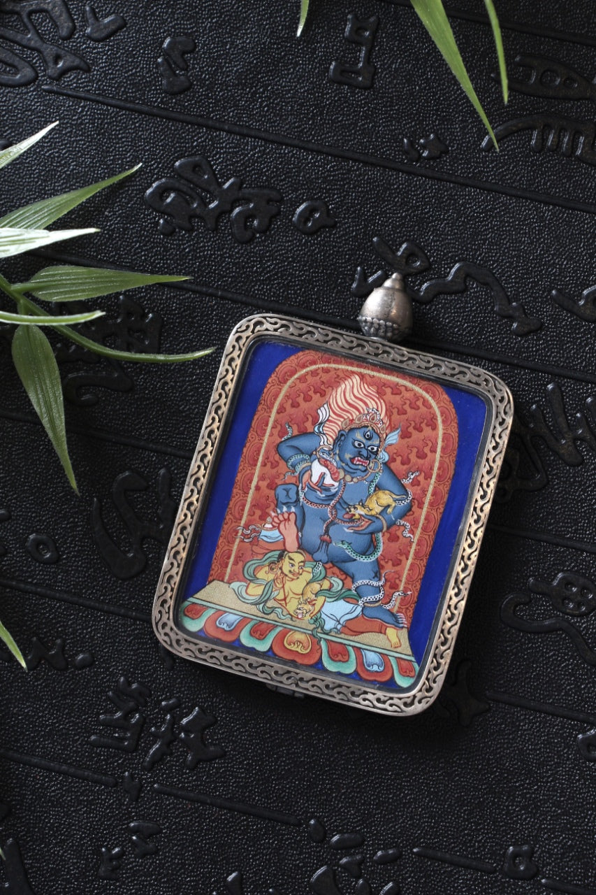 Hand-painted Black Jambhala (Black Wealth Deity) miniature thangka pendant set in a sterling silver gawu case, photographed on a textured black backdrop with bamboo leaves.