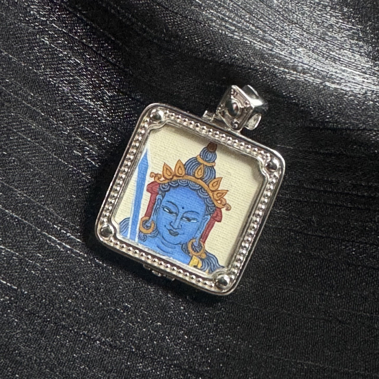 Angled close-up of a square mini thangka pendant showing a single-sided hand-painted blue face with gold crown elements and blue hair details, inside a silver-tone frame with beaded edging and rounded corner beads on black background
