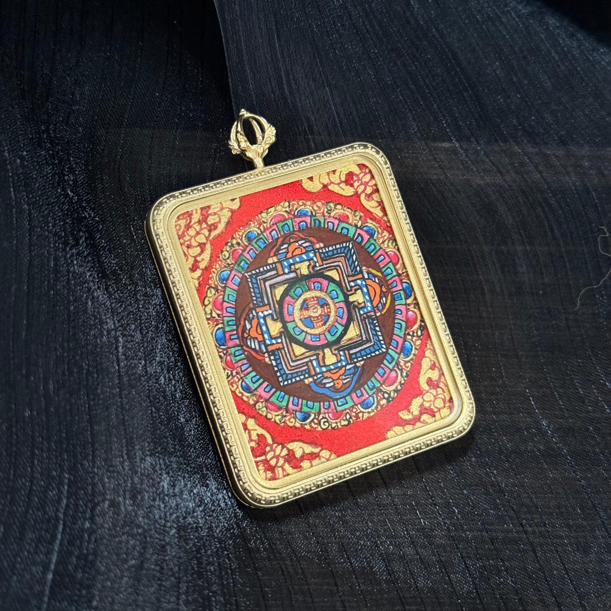 Close view of a Nepalese hand-painted mandala thangka pendant with symmetrical sacred geometry design, vivid mineral pigment colors, and a textured red background inside a gold square frame.