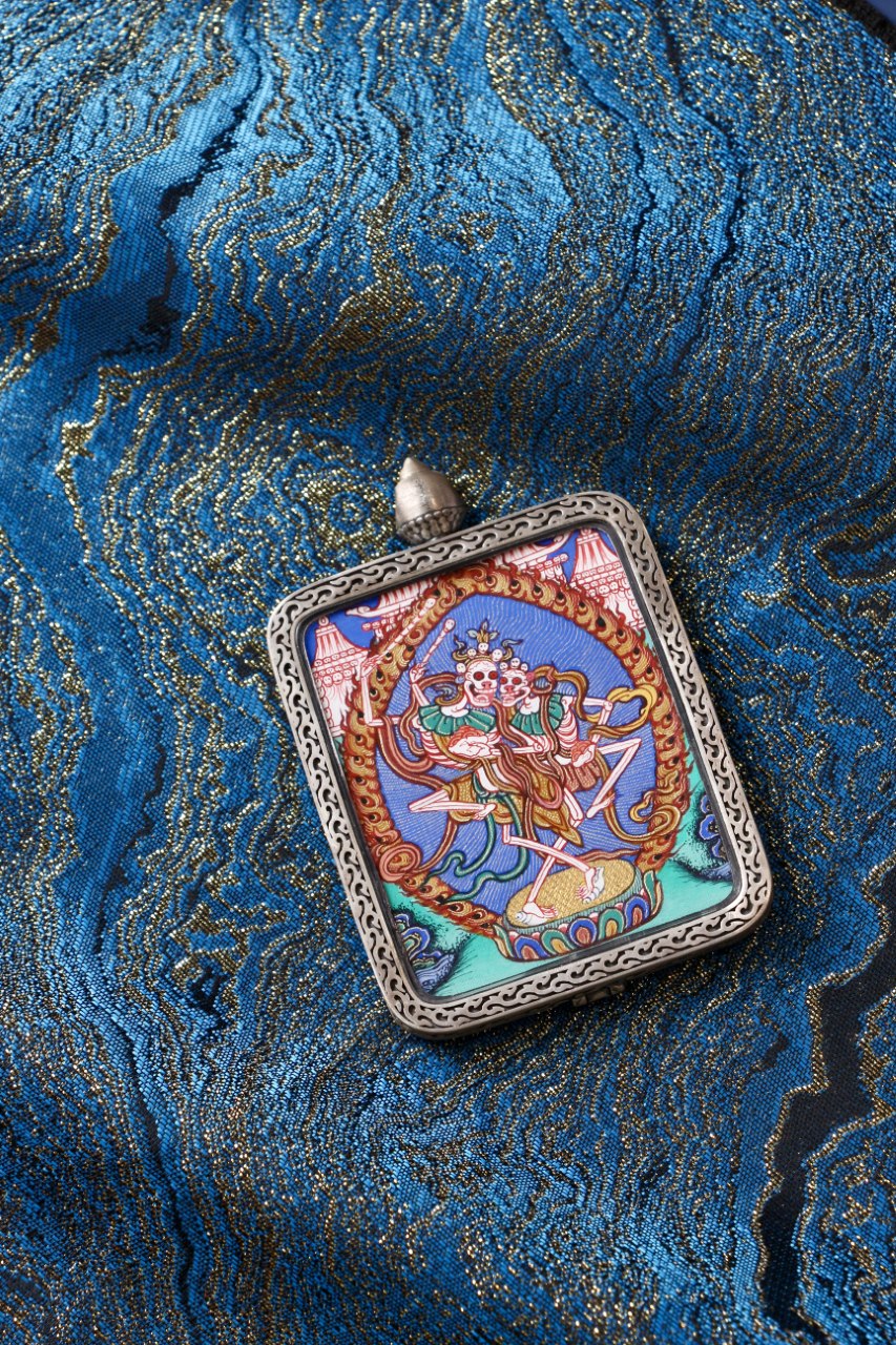 Angled view of Chitipati miniature thangka pendant in silver case on blue brocade background, Regong mineral pigment painting.
