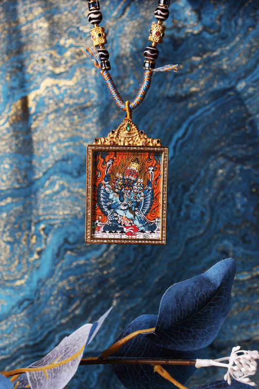 Close-up of a Yamantaka (Vajrabhairava) thangka pendant in a jeweled titanium steel frame, showing detailed deity artwork against a blue-gold textured backdrop.