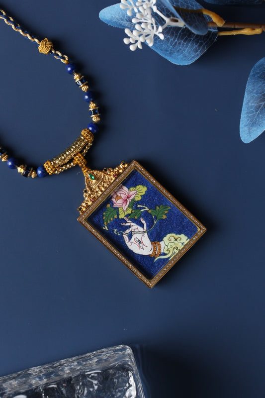 Angled close-up of White Tara hand thangka pendant, lotus flower detail on blue background, gold-tone border with embedded stones, decorative top crown and blue bead necklace strand.