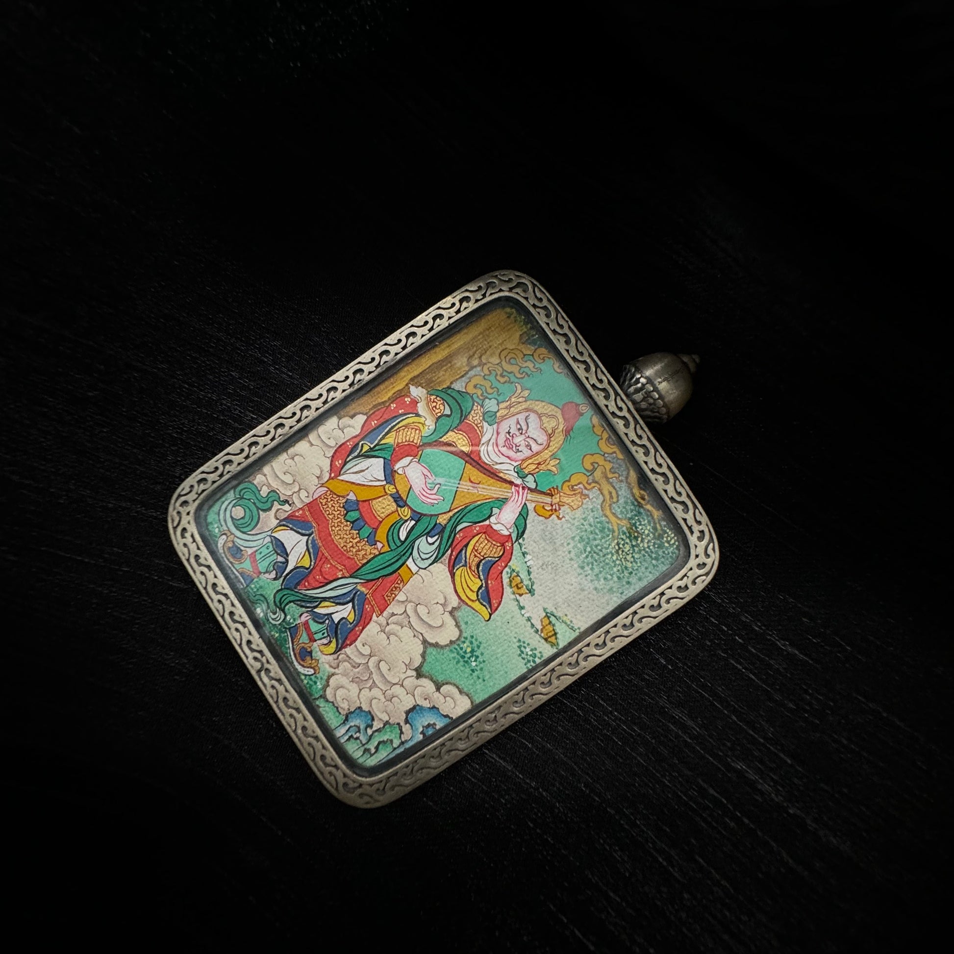 Top view of Tibetan Dhṛtarāṣṭra Eastern Guardian King mini thangka pendant resting on dark fabric, showing hand-painted deity artwork with vibrant colors inside a square sterling silver pendant frame.