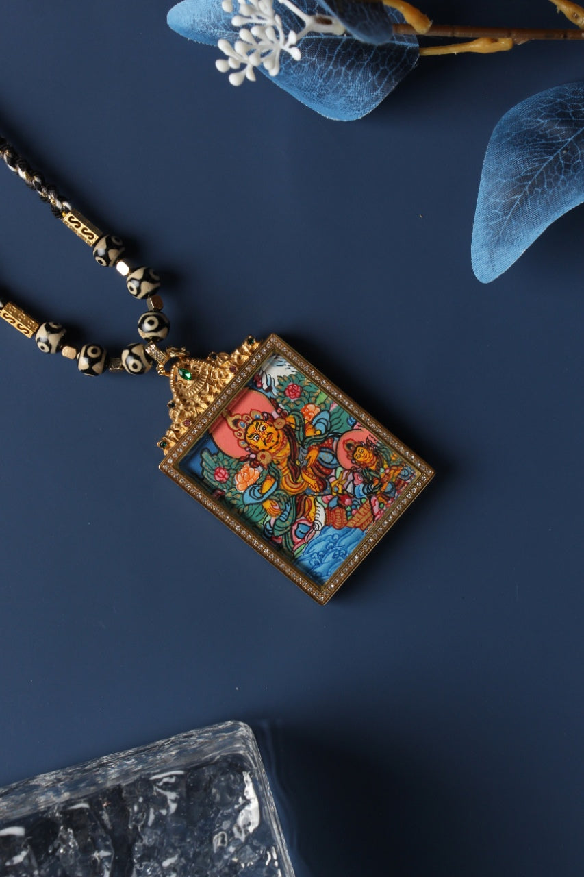 Angled view of the Yellow Jambhala and Vasudhara thangka pendant highlighting the raised metal frame, crystal-set border, and ornate pendant bail with green gemstone.