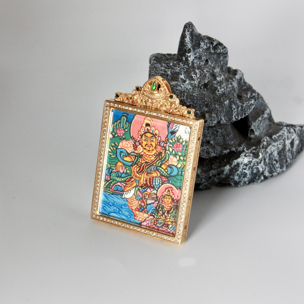 Side angled view of Yellow Jambhala and Vasudhara mini thangka pendant showing raised double-sided titanium steel housing, crystal border, and thickness profile