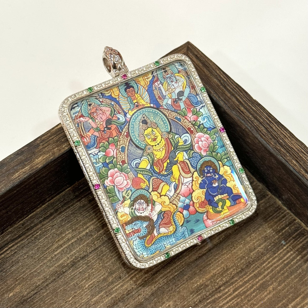 Front scene view of Five Jambhala mini thangka pendant leaning against wooden frames, showing the hand-painted five wealth deities, vivid mineral pigment colors, and the silver gawu-style border with colored accents.