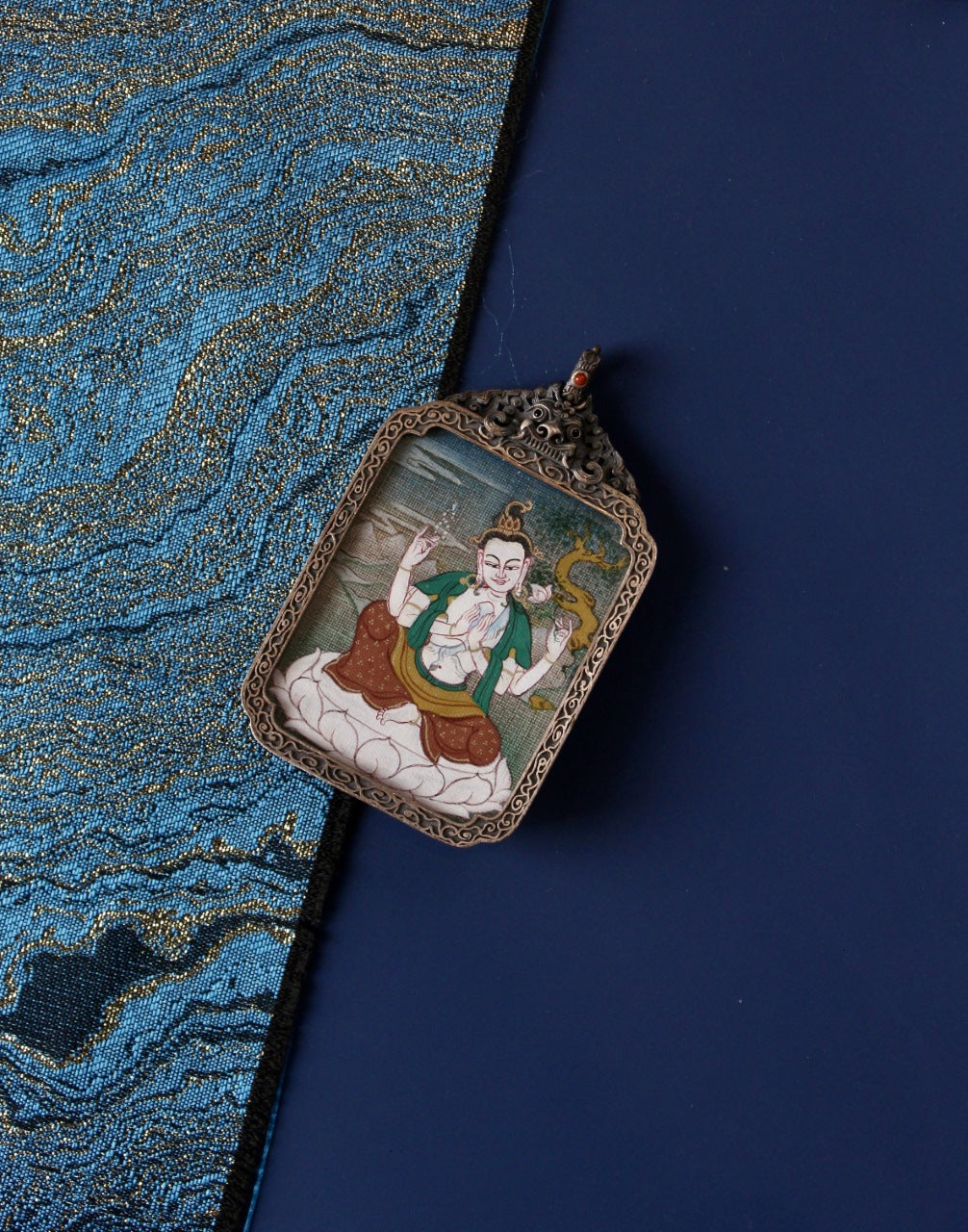 Angled flat lay of Four-Armed Avalokiteshvara mini thangka pendant on traditional textile background, showing the hand-painted thangka artwork in a vintage silver case
