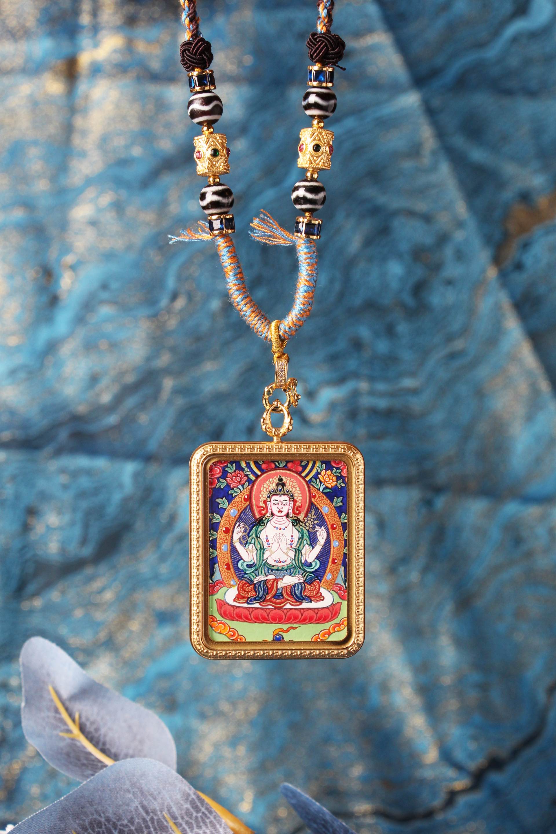 Close-up of the Four-Armed Avalokiteshvara mini thangka pendant showing fine linework, saturated mineral pigment colors, ornate border pattern, and the bead section with metal spacers and braided cord near the pendant loop.