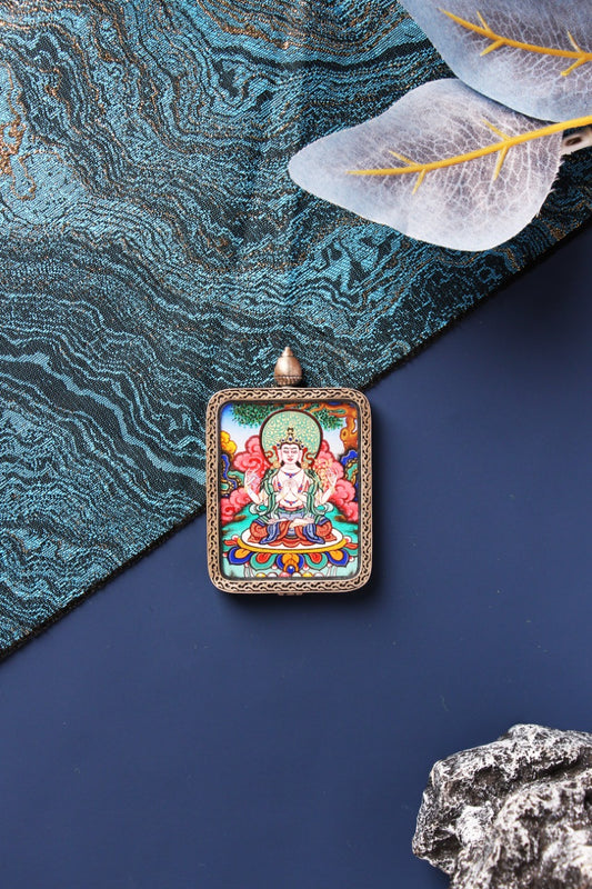 Front view of Four-Armed Avalokiteshvara mini thangka pendant, hand-painted with mineral pigments, featuring the four-armed Chenrezig seated on a lotus base with vivid cloud motifs and a green halo, framed in a Dunhuang Nine-Color Deer-style sterling silver border with a domed top, styled on blue brocade fabric and navy background with translucent leaves.