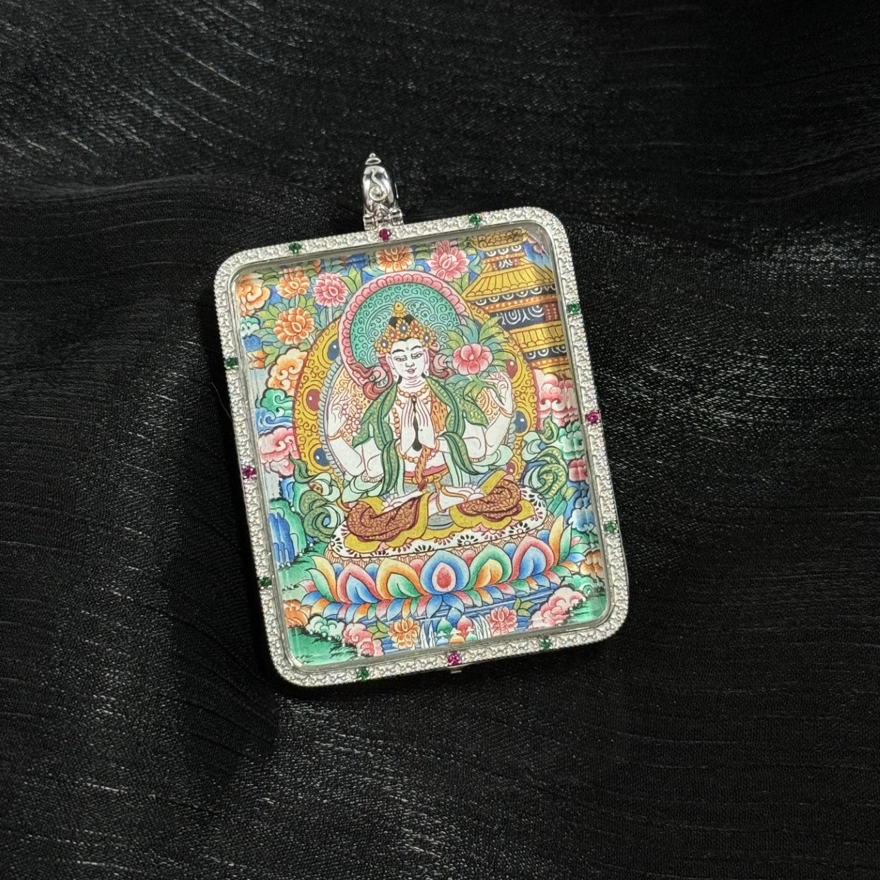 Angled close-up view of a Four-Armed Avalokiteshvara mini thangka pendant highlighting the vibrant mineral pigment painting, lotus base, and ornate Tibetan floral background framed in a 925 sterling silver pendant setting.
