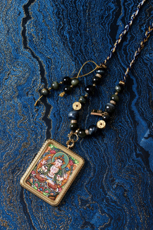 Flat lay of Four-Armed Avalokiteshvara (Chenrezig) hand-painted mini thangka pendant necklace on blue textile background