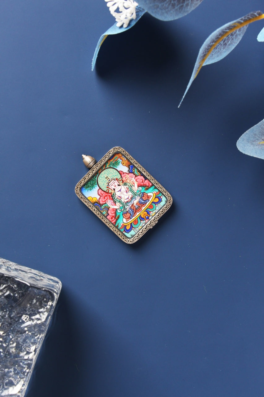Angled flat lay of Four-Armed Avalokiteshvara mini thangka pendant, fully hand-painted with mineral pigments, shown in a dark-toned sterling silver Dunhuang Nine-Color Deer case with patterned edging and a domed top, photographed on a deep navy surface with translucent leaf decor and a clear textured stone prop.