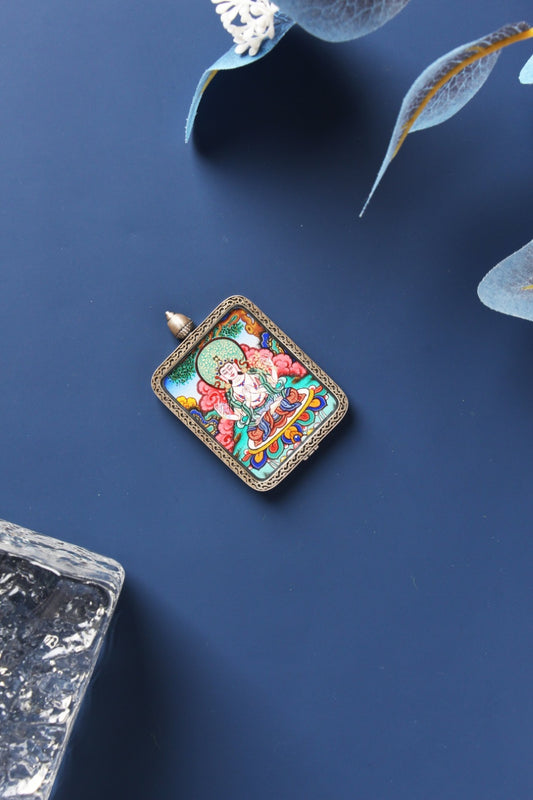 Angled flat lay of Four-Armed Avalokiteshvara mini thangka pendant, fully hand-painted with mineral pigments, shown in a dark-toned sterling silver Dunhuang Nine-Color Deer case with patterned edging and a domed top, photographed on a deep navy surface with translucent leaf decor and a clear textured stone prop.