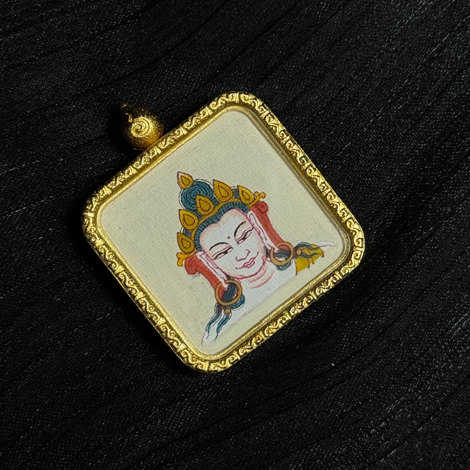 Flat lay view of a Four-Armed Avalokiteshvara thangka pendant placed on dark fabric, revealing the miniature sacred painting on canvas, ornate square gold frame, and traditional Tibetan pendant design suitable for wearable Buddhist devotional art.