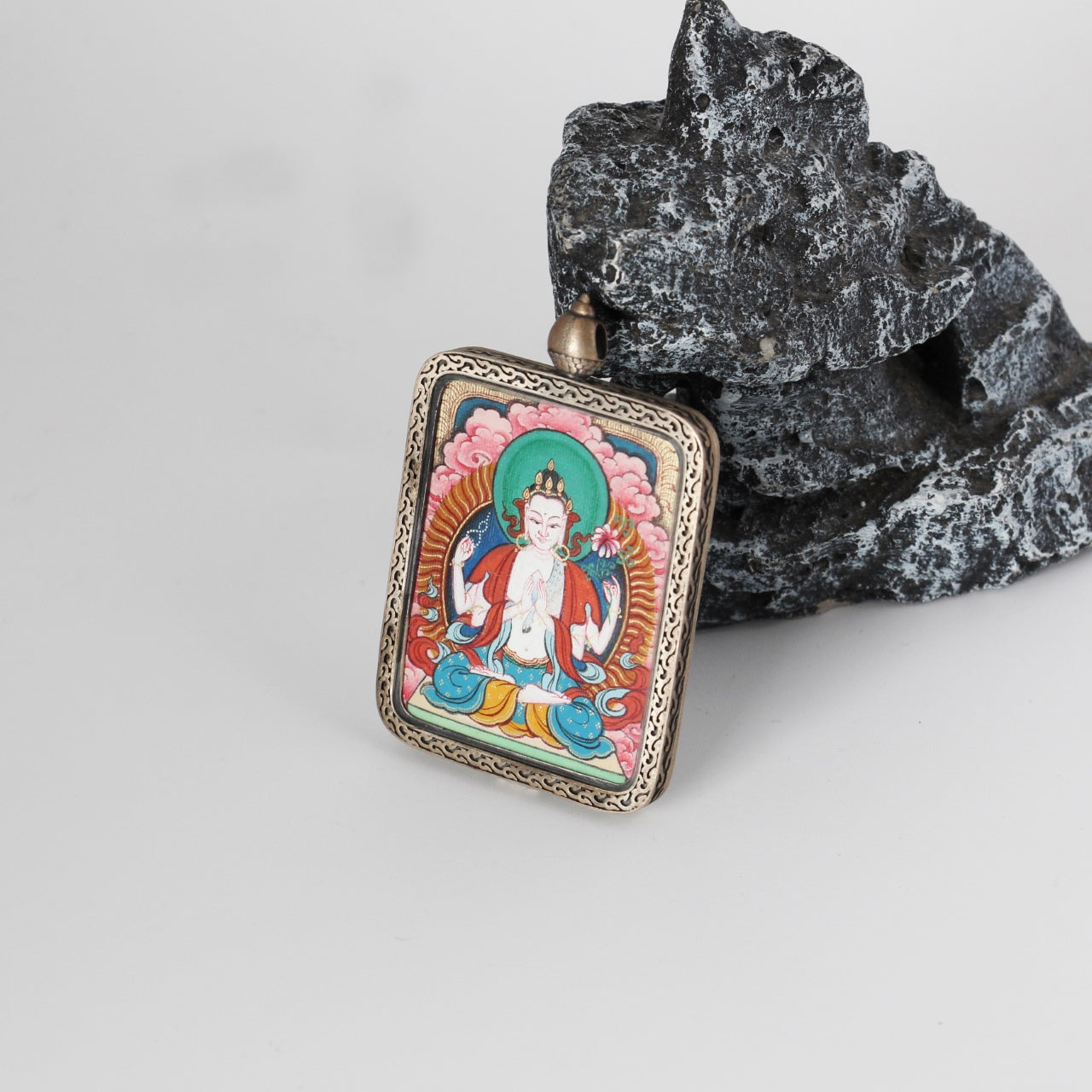 Tilted view of a Four-Armed Avalokiteshvara mini Thangka pendant, showing the engraved silver gawu-style frame, pendant loop structure, and the framed hand-painted Thangka artwork.