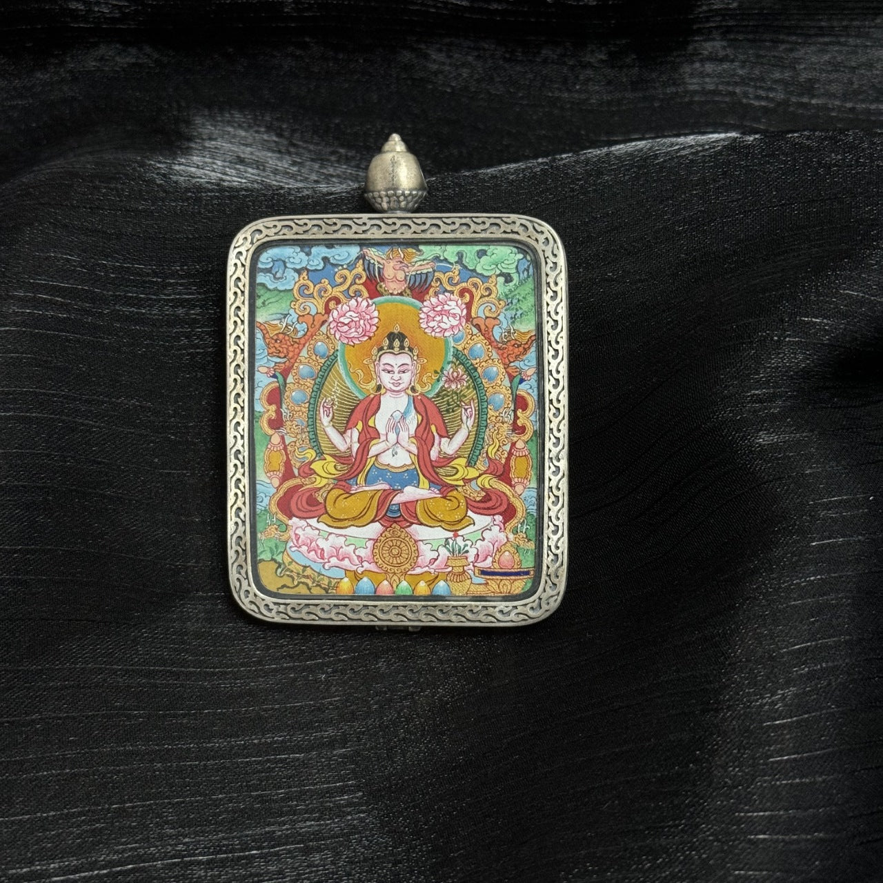 Angled view of Four-Armed Avalokiteshvara mini thangka pendant, hand-painted with mineral pigments, seated on lotus throne with four arms and halo, set in Dunhuang Nine-Colored Deer engraved silver gawu case.