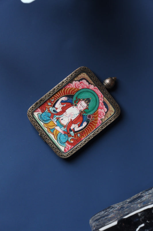 Angled view of a Four-Armed Avalokiteshvara mini thangka pendant, hand-painted with mineral pigments, set in a Dunhuang Nine-Color Deer pure silver gawu-style case.