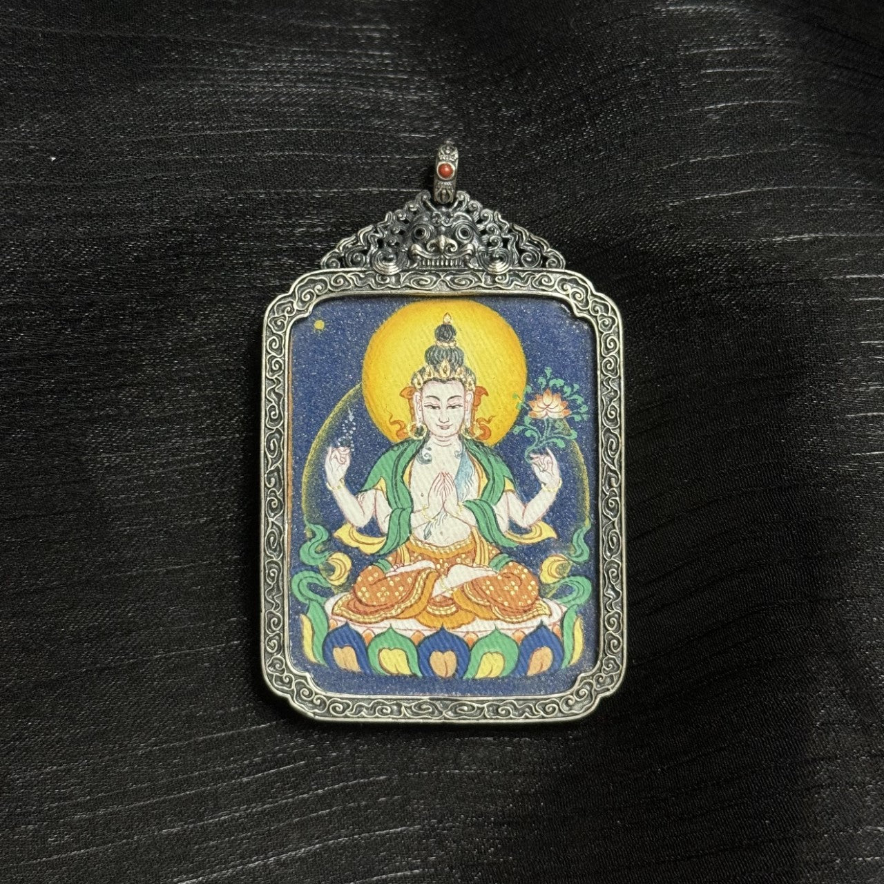 Angled view of Four-Armed Avalokiteshvara mini thangka pendant, hand-painted with natural mineral pigments and set in an ornate vintage silver case with carved decorative patterns.