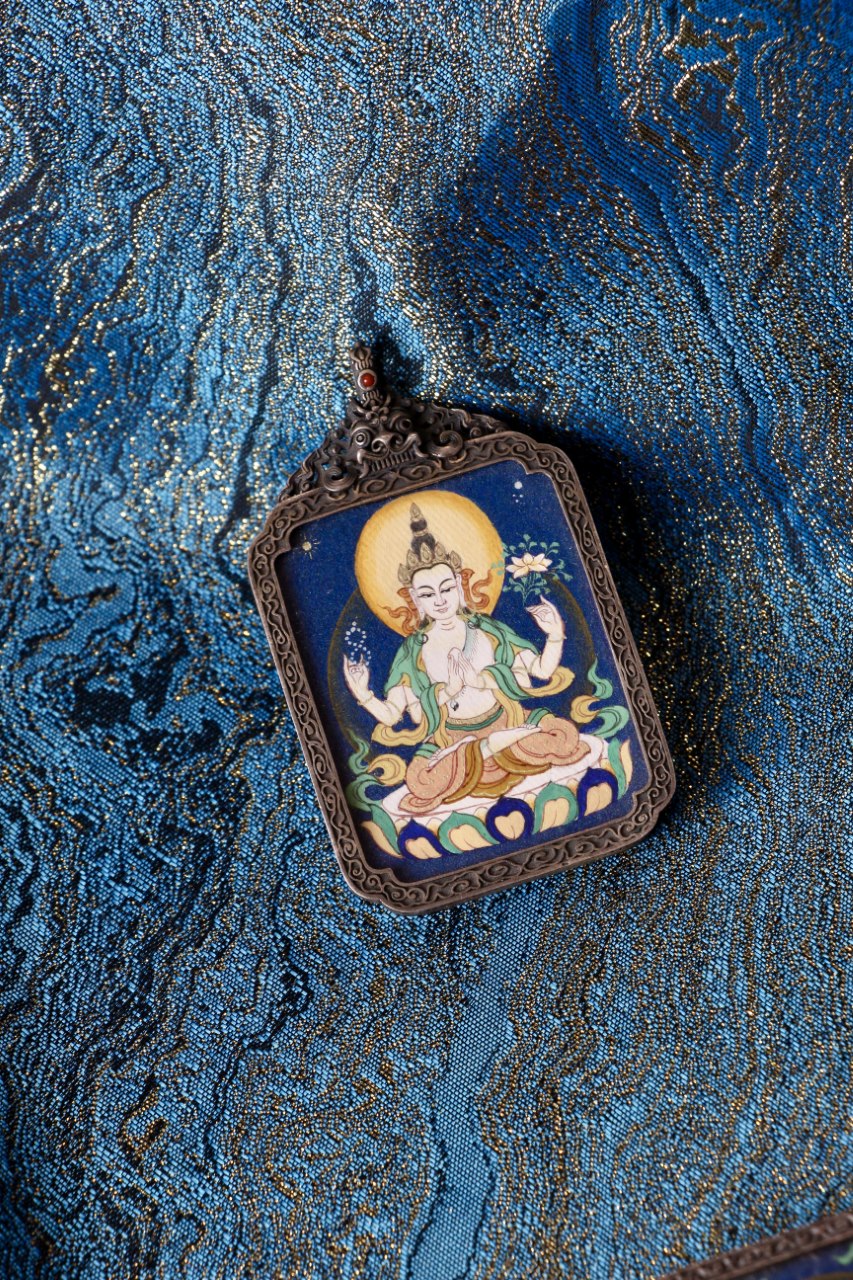 Four-Armed Avalokiteshvara thangka pendant photographed on Tibetan brocade—hand-painted mineral pigments, vintage silver setting; supports calm mind, relieves suffering, and brings peace and wellbeing.