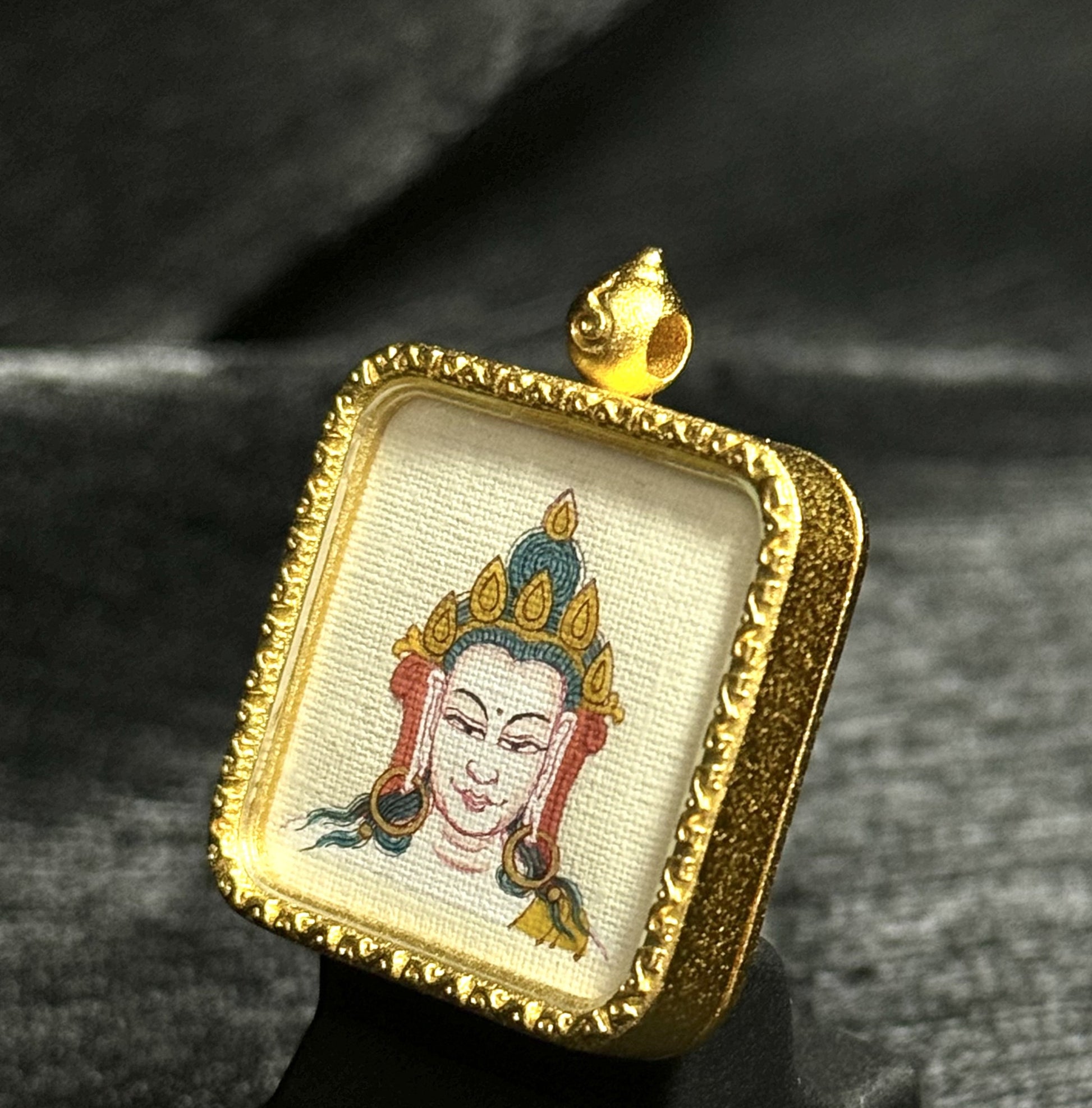 Angled view of a Four-Armed Avalokiteshvara miniature thangka pendant, showing detailed hand-painted deity portrait on textured canvas, natural mineral pigments, and a carved gold-toned square frame with raised ornamental border and hanging loop.